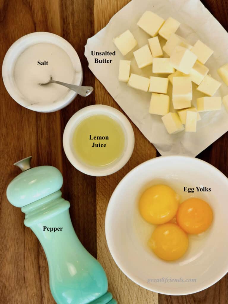 Overhead shot of ingredients for hollandaise sauce, salt, lemon juice, cubed butter, egg yolks, and a pepper grinder.