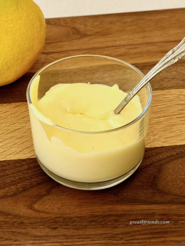 A small glass bowl of hollandaise sauce with a spoon on a wooden board.