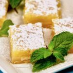 Close up of lemon bars topped with powdered sugar and garnished with a sprig of mint.