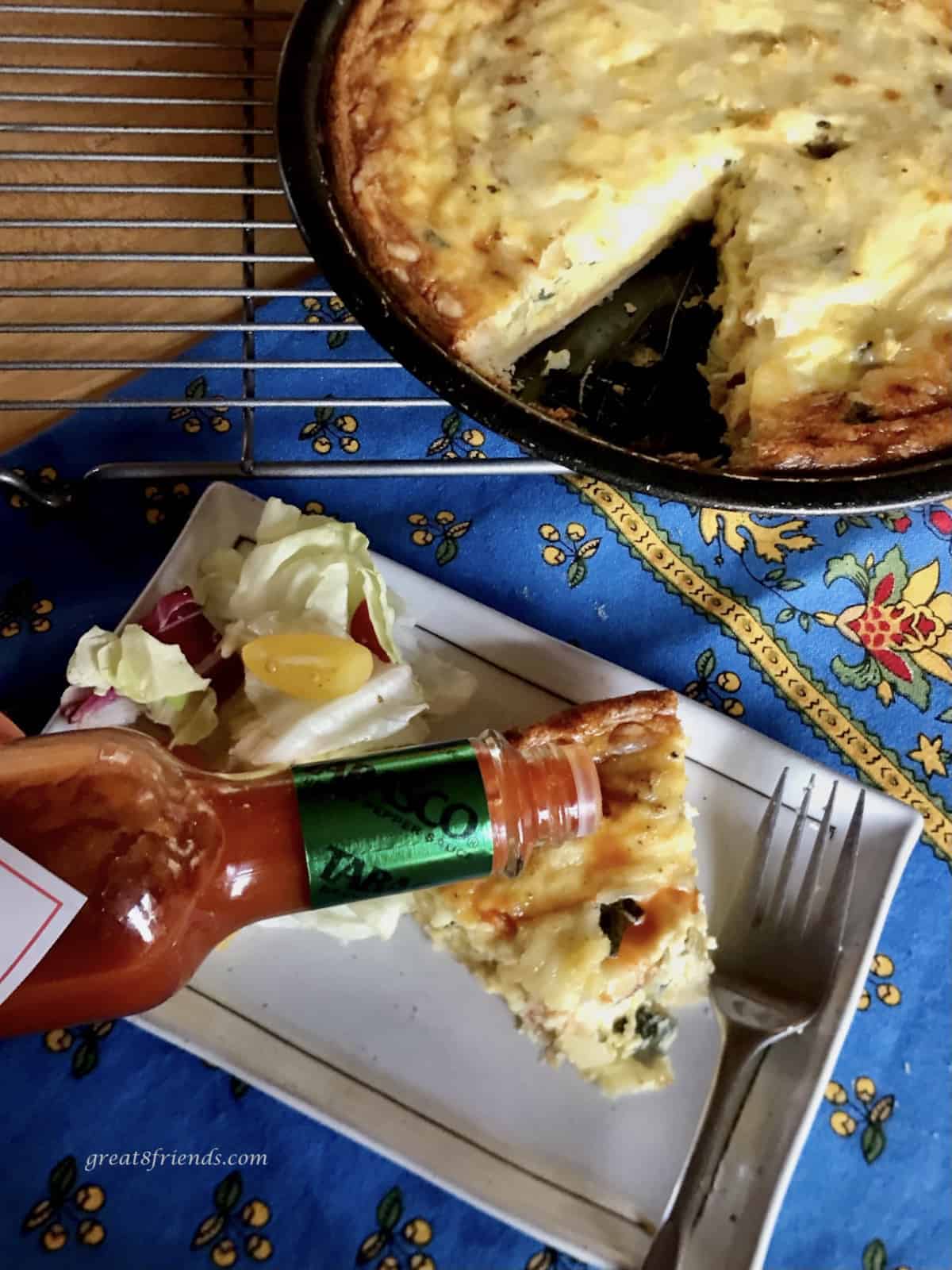 Tabasco hot sauce being added to the top of a piece of quiche on a plate with a salad and the rest of the quiche in a pie plate on the side.