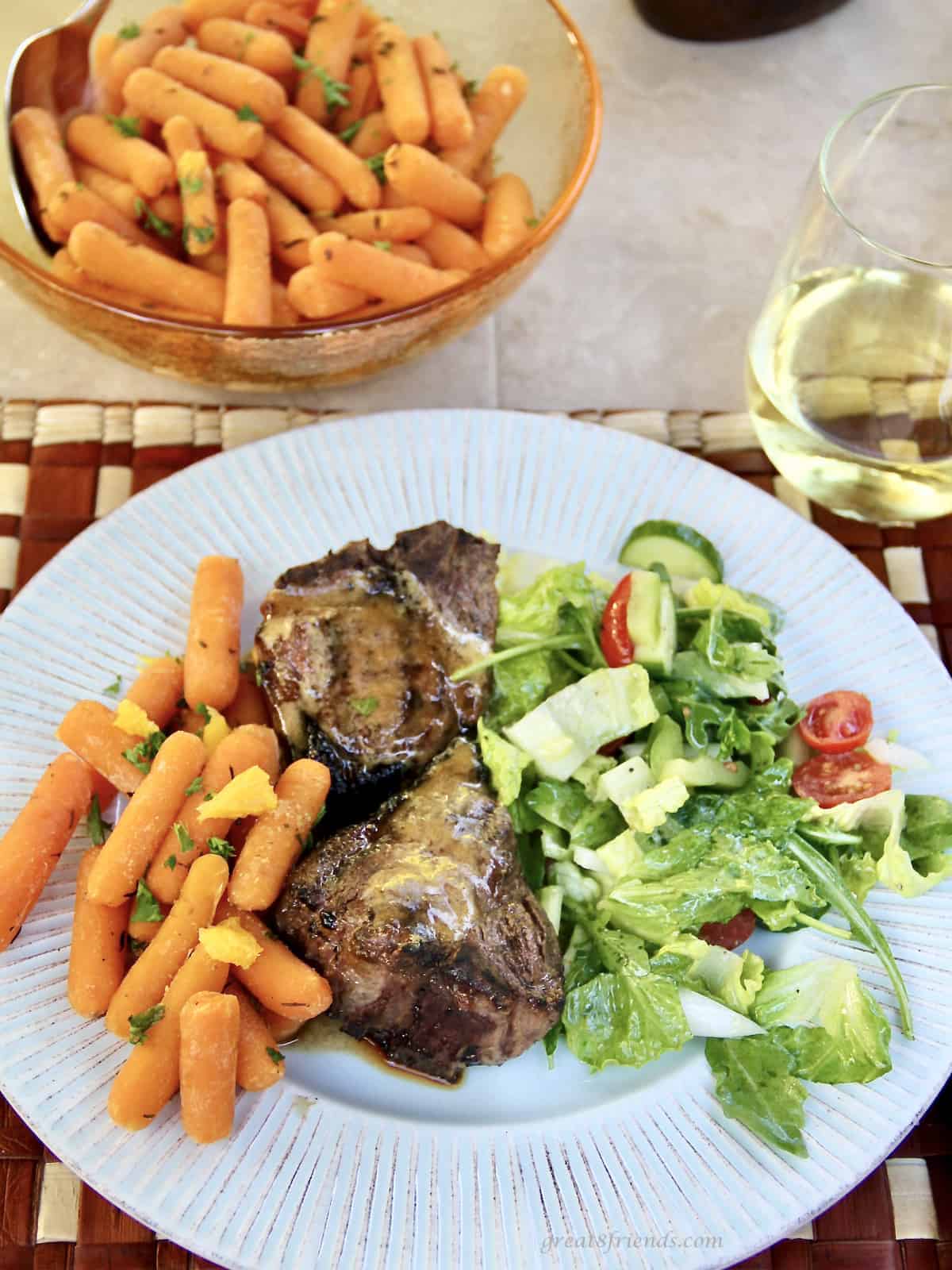 Two grilled lamb chops on a plate with orange baby carrots and a side salad with a bowl of carrots and a glass of white wine on the side.
