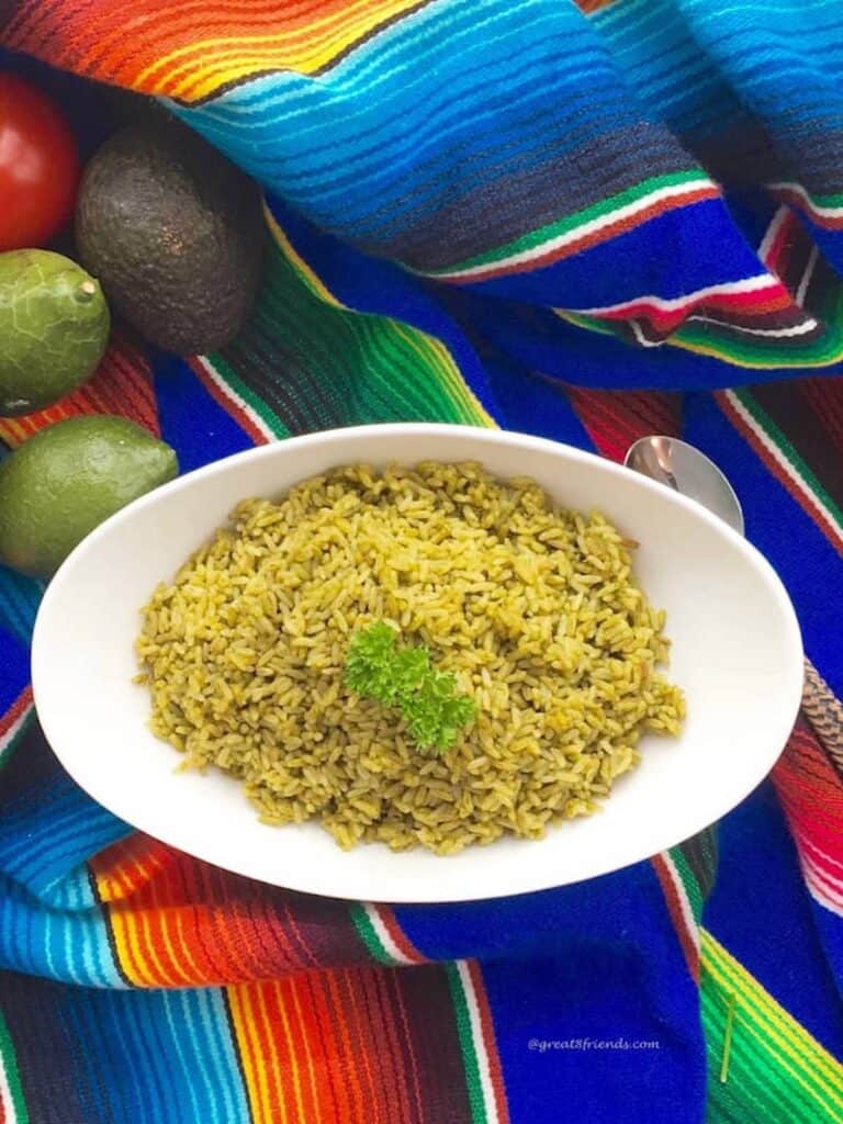 Baked rice with verde ingredients being served in a white oval bowl on a colorful Mexican serape with whole limes on the side.