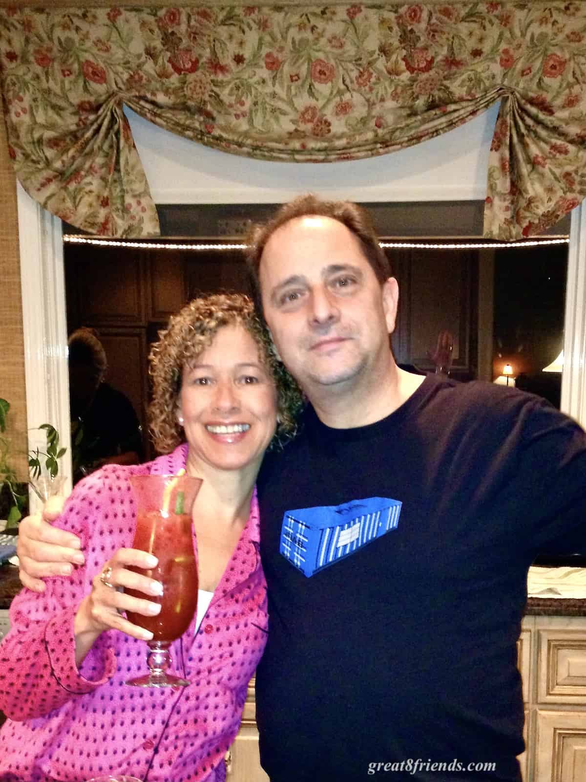 A man and woman posing for the camera and the woman has a tall glass with a Bloody Mary in it.