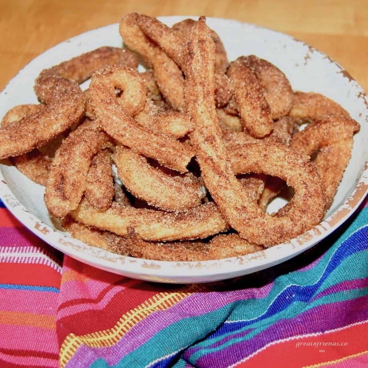 Homemade churros piled on a white plate on a colorful tablecloth.