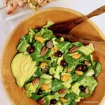 An overhead shot of salad with tangerine slices, pecans, grapes and avocados with wooden salad servers and a tray of grilled shrimp in the background.