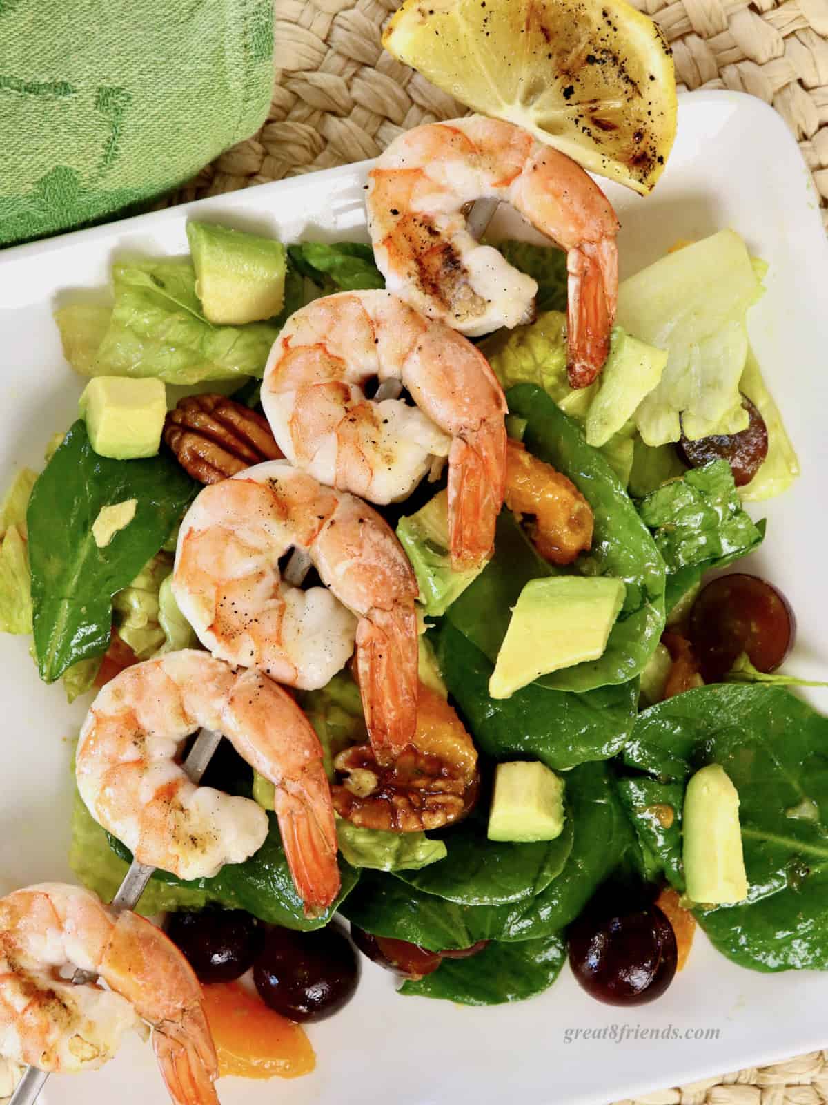 An overhead shot of a serving of salad with tangerine slices, pecans, grapes and avocados and a skewer of grilled shrimp on a square white plate, on a woven placemat.