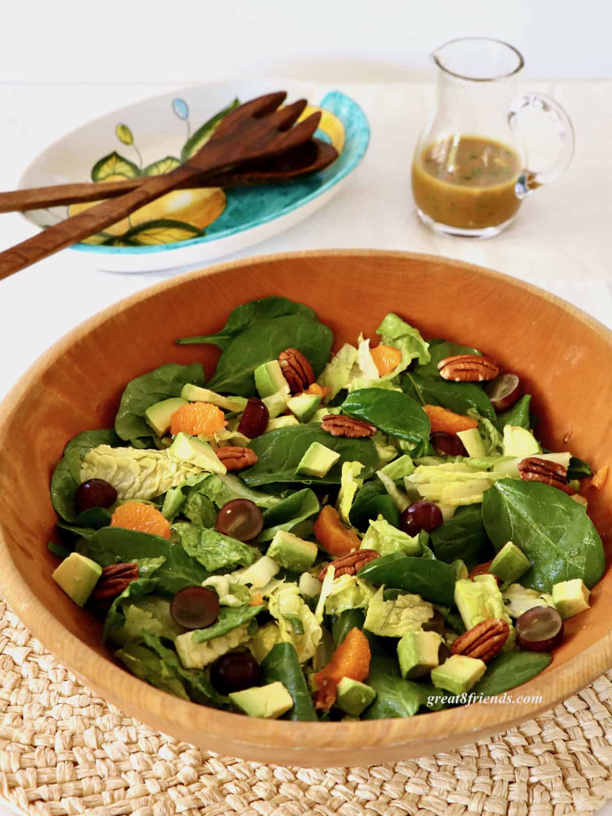 A curried spinach salad with tangerine slices, pecans, grapes and avocados in a wooden bowl with a pitcher of dressing and wooden salad servers in the background.