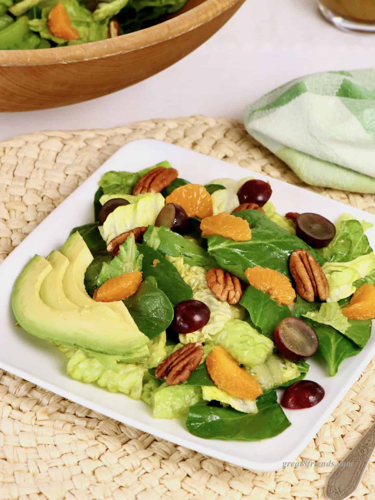 A serving of salad with tangerine slices, pecans, grapes and avocados on a square white plate, on a woven placemat with a green napkin and a wooden bowl of salad in the background.