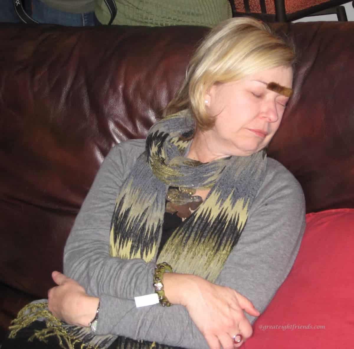 Woman asleep on a couch with a fake mustache on her forehead.