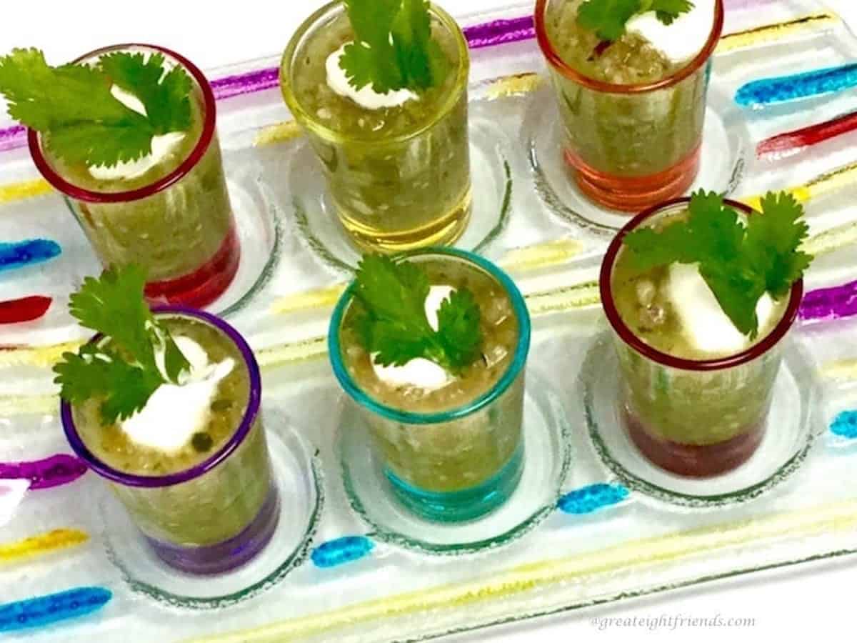 Green gazpacho cold soup being served in six colored glass small shot glasses. 