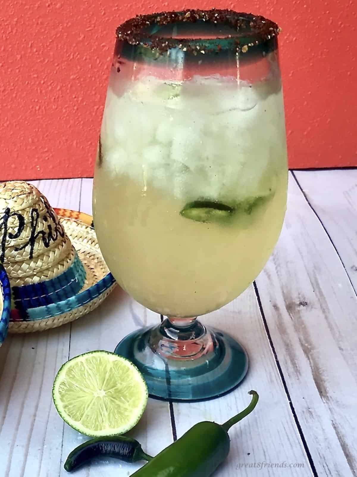 Large glass filledwith ice and a margarita with limes and jalapeños on the table and two mini sombreros with names written on them. 