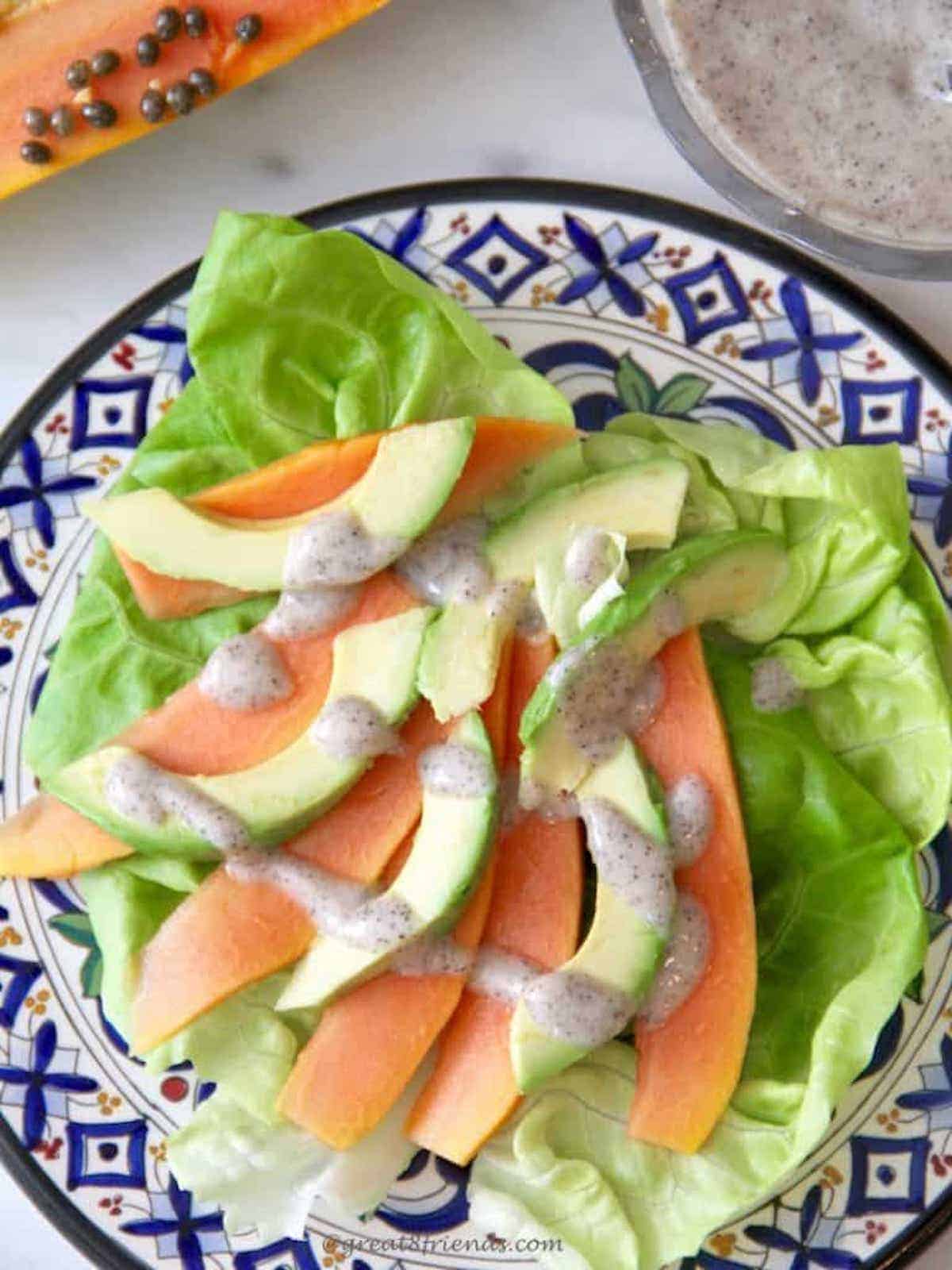 Papaya, Avocado and lettuce salad on a colorful blue plate with a papaya and seeds next to the plate and a bowl of dressing next to the plate.