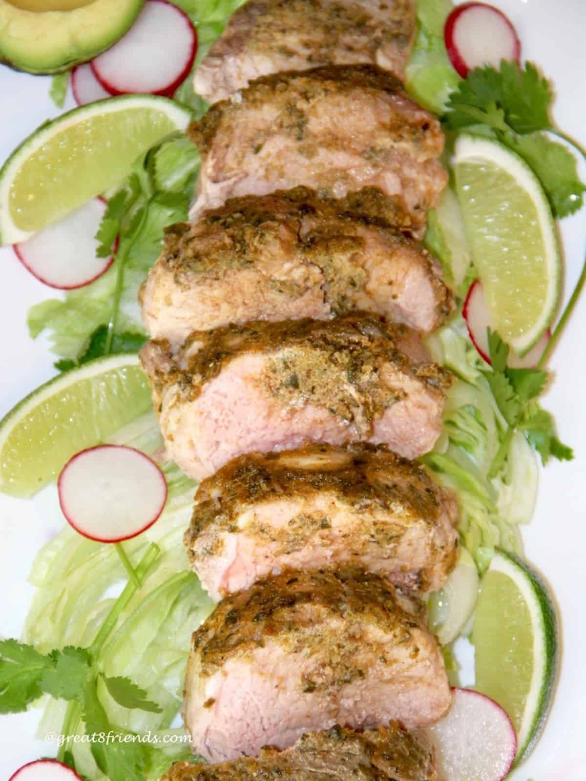 Sliced pork tenderloin on a platter with an adobe sauce on top laying on lettuce, cilantro, limes and sliced radishes.