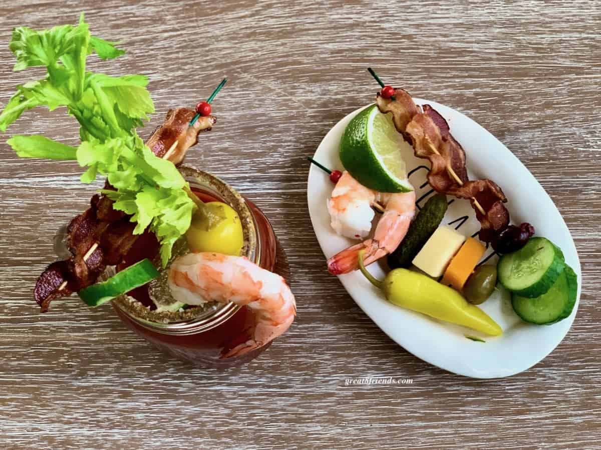 Overhead shot of a white oval plate with a shrimp, slice of lime, skewer of bacon, some cucumbers slices a pickled pepper and some cheese squares alongside a fully loaded bloody mary.