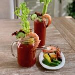 A bloody mary in a glass mug with a shrimp hanging off the side, a slice of cucumber hanging on the side. There's another bloody Mary in the background along with a small plate of extra garnishes.