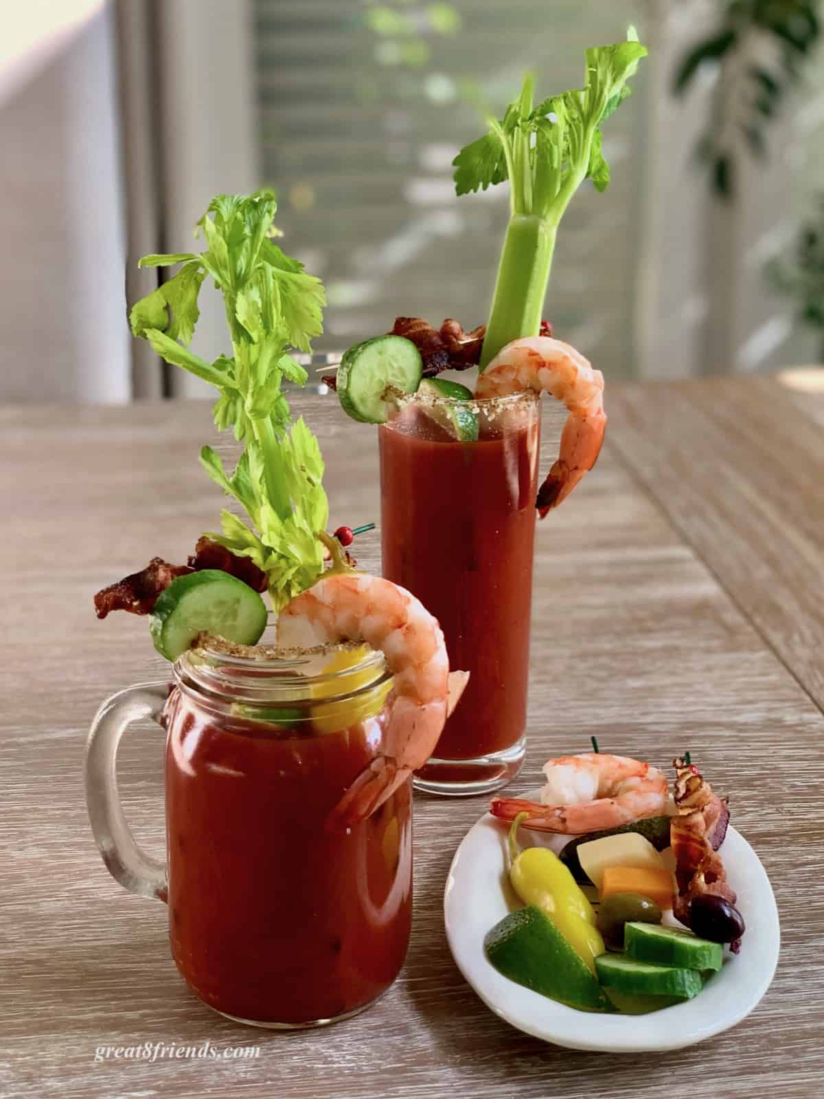 A bloody mary in a glass mug with a shrimp hanging off the side, a slice of cucumber hanging on the side. There's another bloody Mary in the background along with a small plate of extra garnishes.