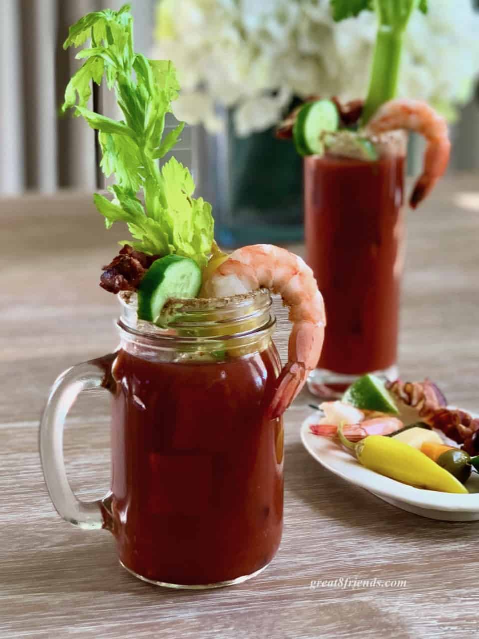 A bloody mary in a glass mug with a shrimp hanging off the side, a slice of cucumber hanging on the side. There's another bloody Mary in the background along with a small plate of extra garnishes.