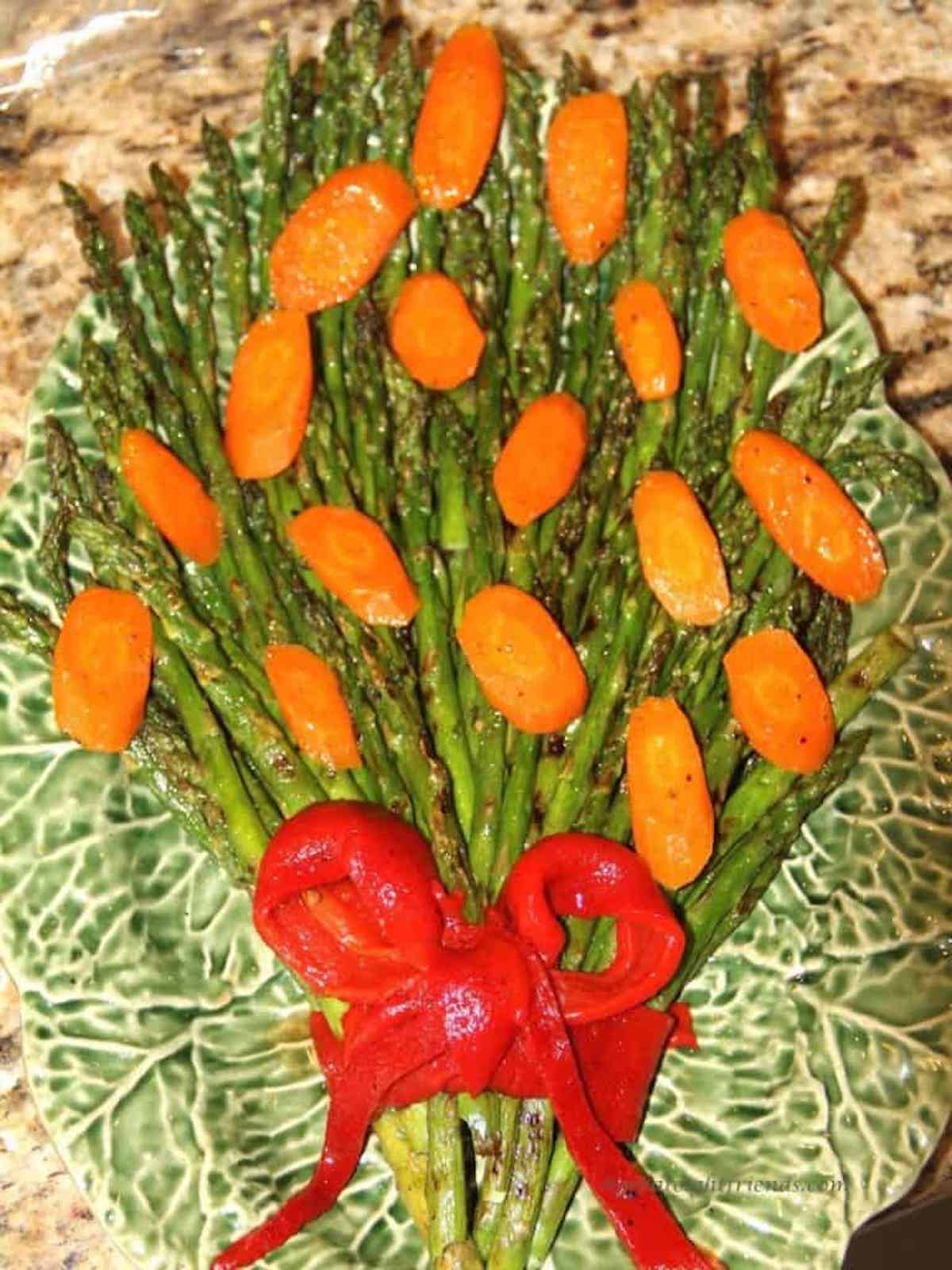 Large green ceramic platter with asparagus spears laid on it in a bouquet form with cut carrots as the flowers and red pepper strips as the bow.