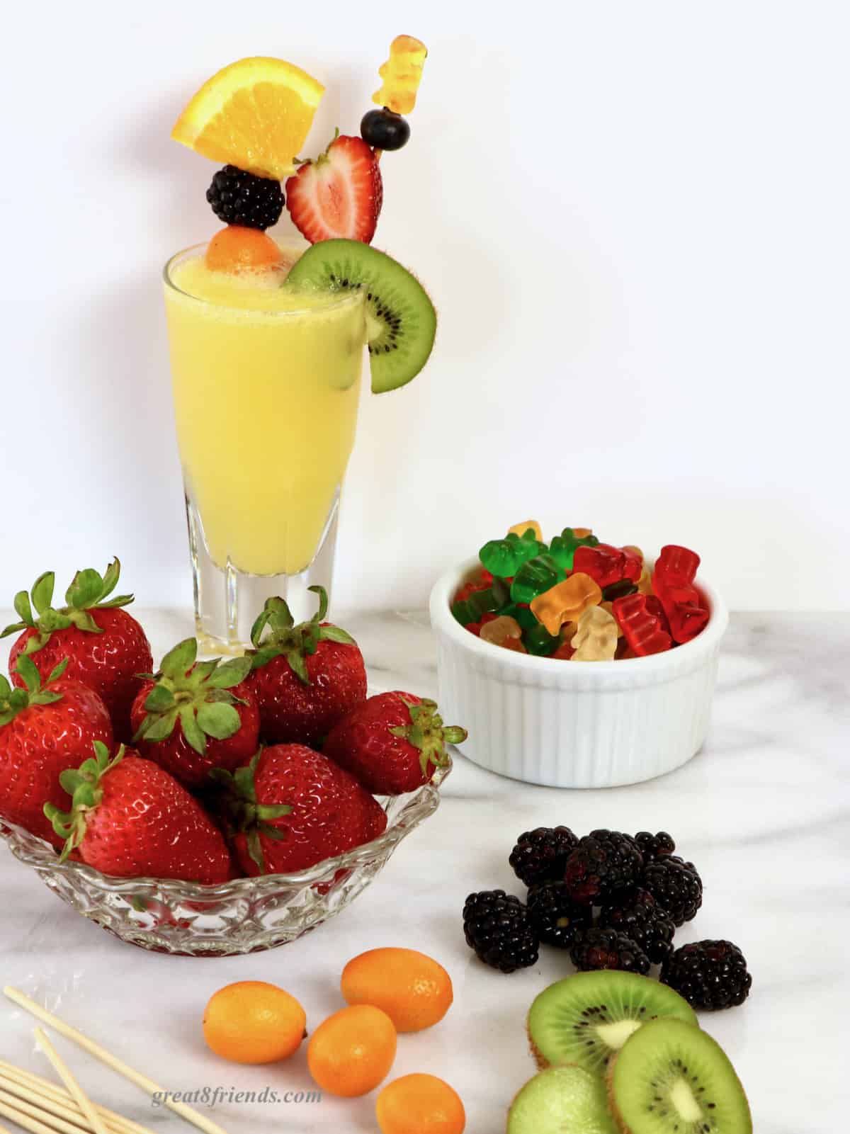 A mimosa in a juice glass garnished with two skewers of assorted fruit on marble with a bowl of strawberries, a bowl of gummy bears, some kumquats, blackberries, and kiwifruit.