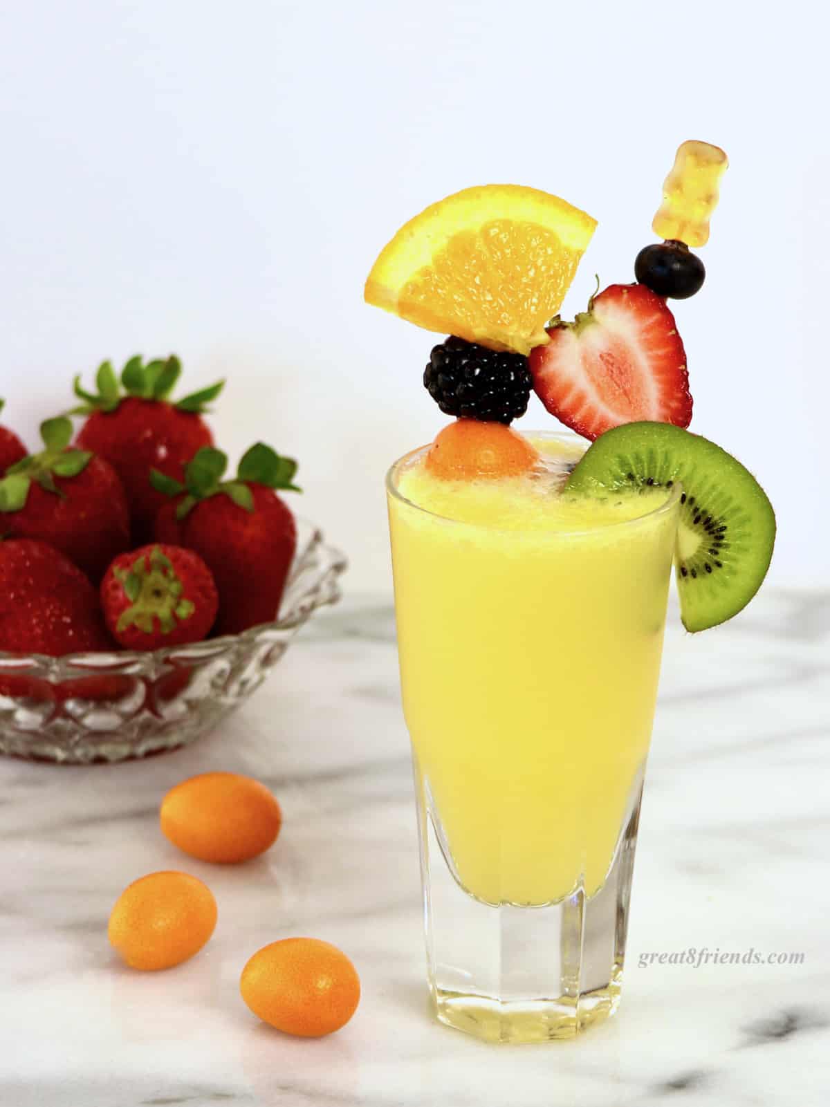 A mimosa garnished with 2 skewers of fresh fruit with a bowl of strawberries and some kumquats in the background.