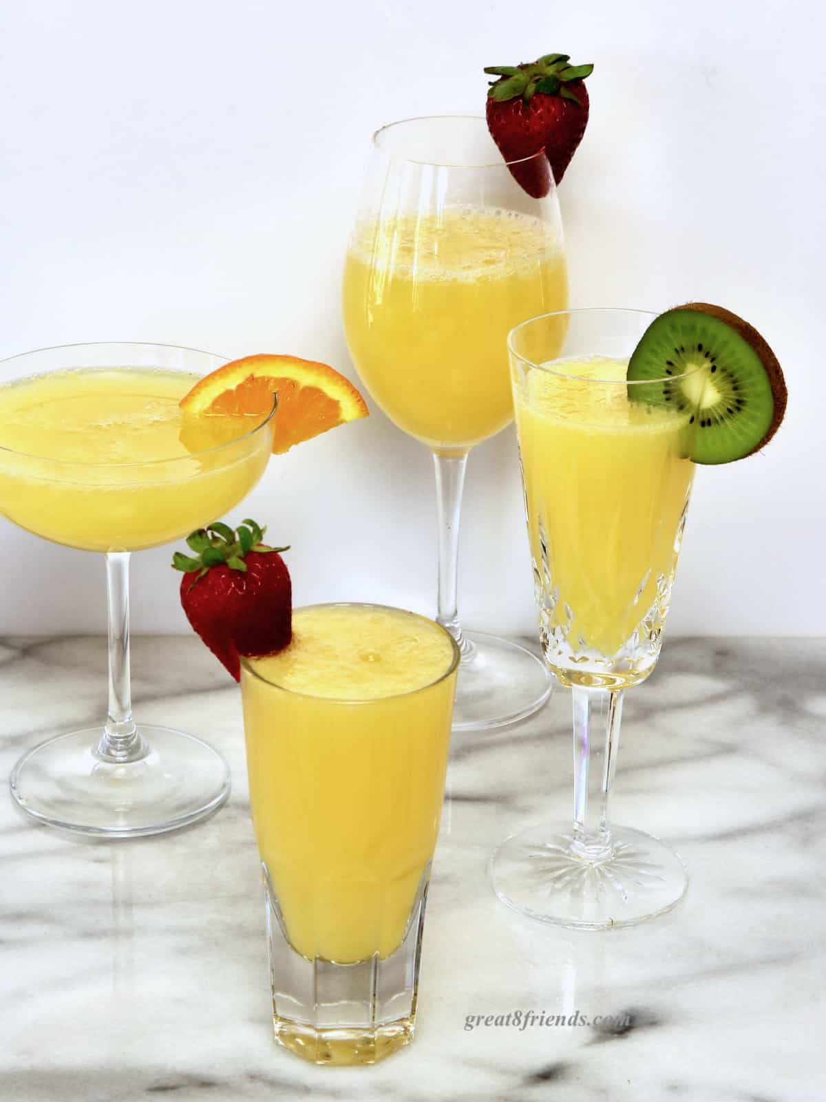 Four mimosas each in a different glass; one wine glass garnished with a strawberry, one coupe glass garnished with an orange slice, one flute garnished with a slice of kiwi, one juice glass garnished with a strawberry.